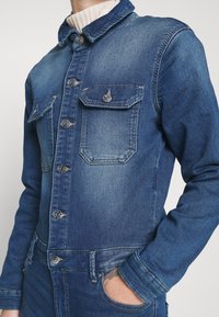 Homme portant une veste en jean bleue et un jean assorti, main dans la poche, montrant les détails des boutons et les poches poitrine à rabats.