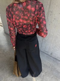 Rot und schwarz gemusterter Langarmoberteil mit Kirschmuster; kombiniert mit schwarzen, weit geschnittenen Hosen mit Gesäßtaschen und einem roten Logo, während eine braune Tasche getragen wird.