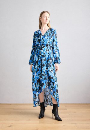 Robe portefeuille bleue à motifs floraux avec manches longues, fente haute et fond noir. Portée avec des bottines à talons aiguilles noires. Tissu doux.