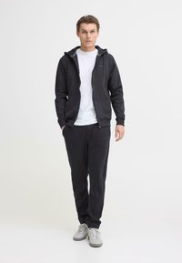 Ciemnoszara bluza rozpinana na zamek z przednimi kieszeniami, zestawiona z dopasowanymi dresami i białym t-shirtem. Gładka faktura, elastyczne mankiety i luźny krój.