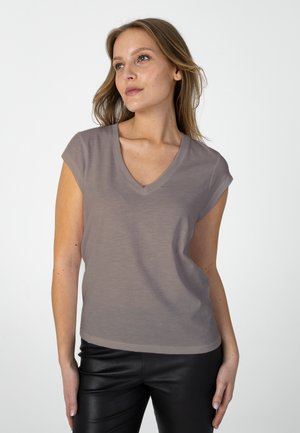 Woman with shoulder-length hair wearing a taupe short-sleeve V-neck top and black pants, looking slightly upward to the left.