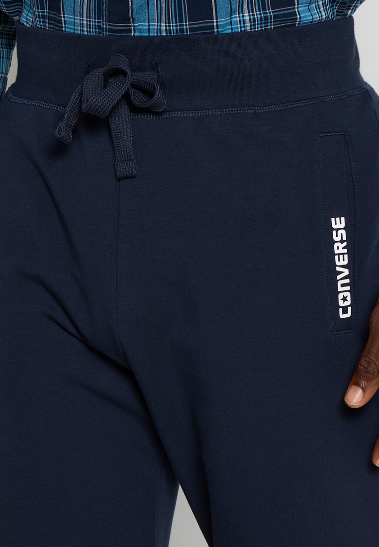 Pantaloni jogger blu navy con vita regolabile con cordino, tasca laterale e logo "Converse" bianco. Realizzati in morbido cotone, presentano una texture liscia.