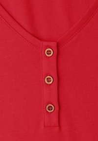 Tissu rouge avec trois boutons beige alignés verticalement sur une patte sous un col arrondi, montrant des coutures détaillées et la texture.
