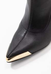 Czarne skórzane botki z ostrym noskiem, z złotym metalowym czubkiem, na którym jest wygrawerowane "Versace Jeans Couture", na białym tle.