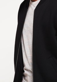 Schwarze Bomberjacke aus glattem Material, mit Reißverschluss vorne und geripptem Kragen, getragen über einem hellgrauen T-Shirt.