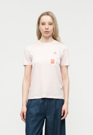 Νεαρή γυναίκα με ξανθά μαλλιά φοράει ανοιχτόροζ μπλουζάκι με τσέπη στο μπροστινό μέρος που έχει κόκκινα φρούτα ως σχέδιο και σκούρο μπλε παντελόνι.