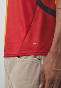 Rød og orange performance trøje med mesh struktur, der har en diagonal sort detalje og "DRYCELL" label i sømmen.