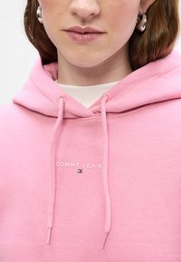 Sudadera con capucha rosa y cordones, con el logo "TOMMY JEAN" y un pequeño emblema de la marca. Textura de tela suave.