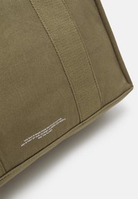 Zbliżenie na róg oliwkowej torby z materiału, z wzmocnionym przeszyciem i białym napisem informującym, że została wykonana z recyklingowanej bawełny.