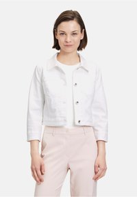 Weiße Denim-Cropped-Jacke mit langen Ärmeln, zwei Brusttaschen und vorderem Knopfverschluss. Trägt sie über einem weißen Oberteil und hellrosa Hose.