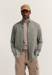 Mann mit beigem Hut, hellgrauem Rollkragenpullover, offenem grauem Hemd und beigen Cargo-Hosen, mit den Händen in den Taschen vor einem schlichten Hintergrund.