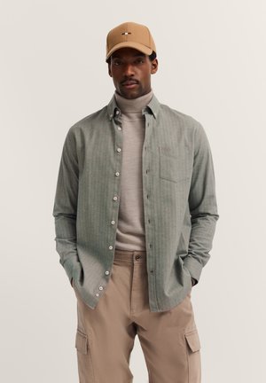 Man wearing beige cap, light gray turtleneck, unbuttoned gray shirt, and beige cargo pants with hands in pockets against plain background.