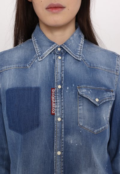 Light blue denim shirt featuring a pointed collar, two chest pockets, red logo tag, and noticeable fading and distressing details.
