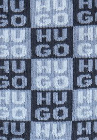 Dzianinowa tkanina z wzorem w kratkę, składająca się z naprzemiennie ułożonych kwadratów w kolorze granatowym i jasnoniebieskim. Na tkaninie widnieje napis „HUGO” w odważnym, powtarzającym się wzorze.