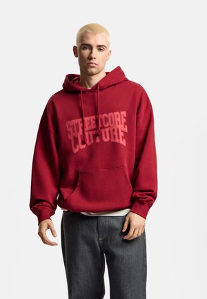 Młoda osoba z krótkimi blond włosami, ubrana w czerwoną bluzę z kapturem z napisem "STREETCORE CULTURE" oraz ciemne dżinsy na jednolitym tle.