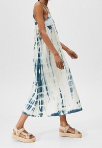 Donna che cammina con un abito midi senza maniche blu e bianco tie-dye e sandali con plateau beige su uno sfondo semplice.