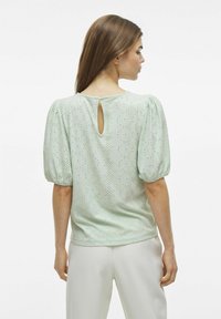Blusa verde menta con maniche a sbuffo corte, dettaglio a chiave sulla schiena e motivo ad occhiello su tutto il tessuto. Materiale morbido e testurizzato.