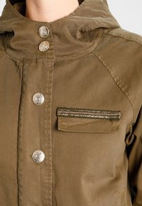 Beige jacka med huva, med metallicknappar, en bröstficka med texturerad kant och en slät tygfinish.