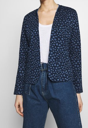 Woman wearing a navy blue patterned blazer with a white shirt and high-waisted blue jeans.