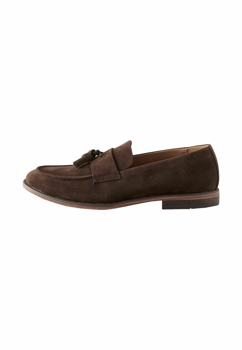 Mocasines de ante marrón con una correa decorativa y detalle de borlas, punta redondeada y un tacón bajo y apilado. Textura suave y diseño sencillo.