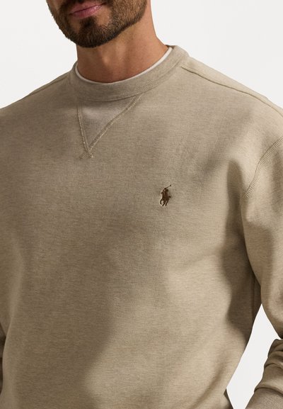 Lys beige genser med rund hals laget av myk bomullsblanding, med en liten brodert logo på venstre bryst og ribbestrikkede mansjetter.