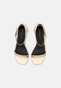 Beige hælhæle sandaler med ankelrem, glat tekstur, firkantet tå og sort indvendig foring; har guldspænde som pynt.