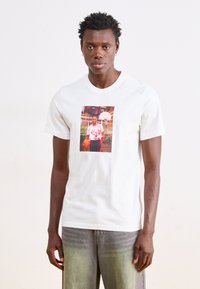 Λευκό βαμβακερό μπλουζάκι με πολύχρωμο εκτυπωμένο γραφικό σχεδίασης ενός καλαθοσφαιριστή, στρογγυλή λαιμόκοψη, κοντά μανίκια και χαλαρή εφαρμογή.