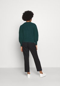 Ciemnozielony sweter dzianinowy z okrągłym dekoltem i bufiastymi rękawami, w połączeniu z czarnymi zwężanymi spodniami i białymi sneakersami.