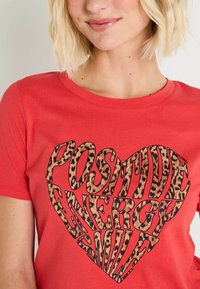 Camiseta roja de manga corta con un diseño de corazón de estampado de leopardo y un texto en negrita negro que dice "SONRISA ENERGÍA POSITIVA". Textura de tela suave.
