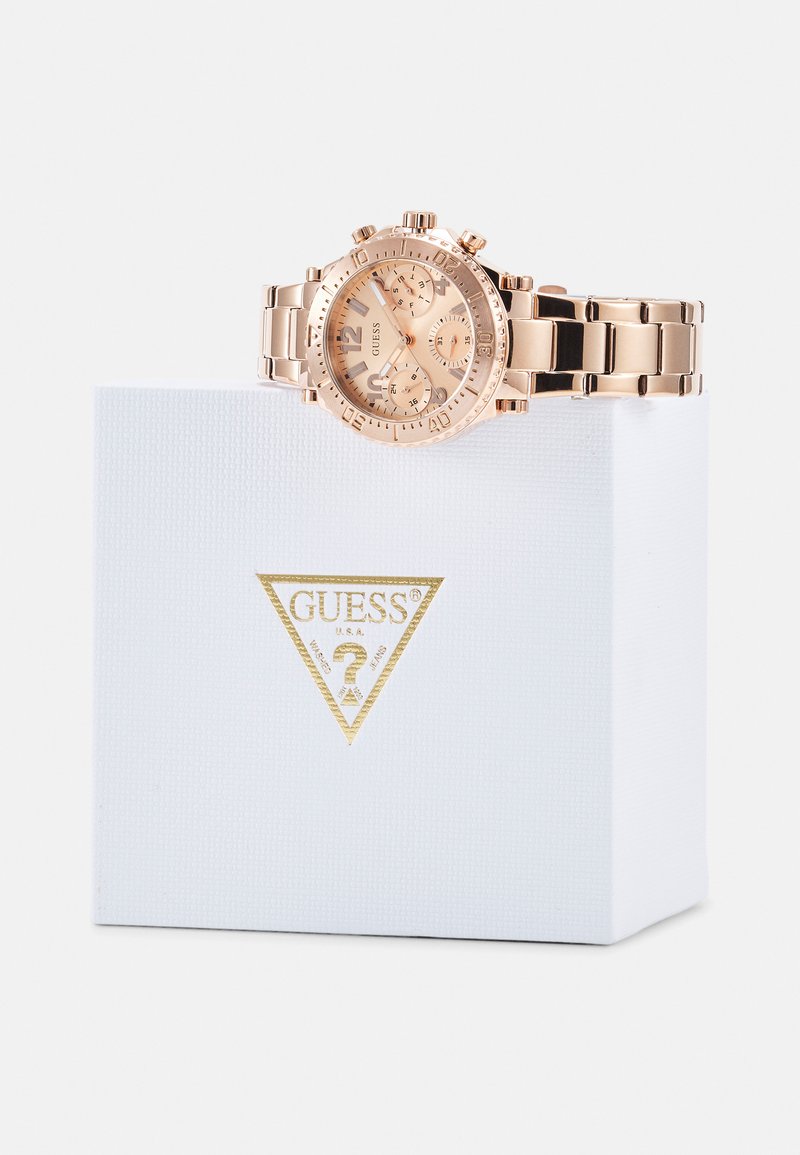 Montre chronographe en or rose avec trois sous-cadrans, bracelet en métal et lunette texturée, placée sur une boîte blanche marquée GUESS.
