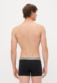 Jovem visto de costas usando cuecas boxer pretas da Tommy Hilfiger com cintura cinzenta, contra um fundo branco simples.
