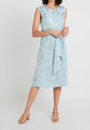 Vrouw draagt een mouwloze lichtblauwe bloemen midi-jurk met een gestrikte tailleband en beige sandalen met hak, staand tegen een effen achtergrond.