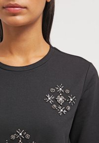Sudadera negra con cuello adornado que presenta acentos de perlas y cuentas plateadas en diseños florales y geométricos. Textura de tela suave.