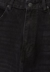 Gros plan sur l'avant d'un jean en denim noir avec bouton, passants de ceinture, bords de poches et détails de coutures visibles.