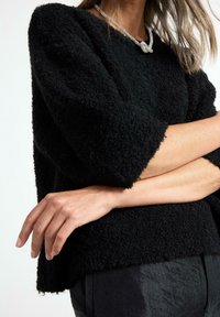 Musta tekstuuriga sviiter, millel on vabalt langev lõige ja kolmveerandi pikkuses varrukad. Selle alla on kantud tume püksid, millel on peen sära.