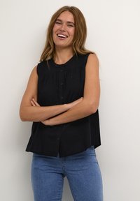 Blusa nera senza maniche con chiusure a bottone, frontale plissettato e colletto arrotondato. Abbinata a jeans aderenti blu chiaro.