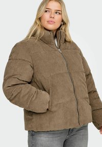 Smeđa pufasta jakna od korduroja s visokim ovratnikom, prednjim patentnim zatvaračem i bočnim džepovima; s rebrastom teksturom i detaljima prošivenim blazinicama.