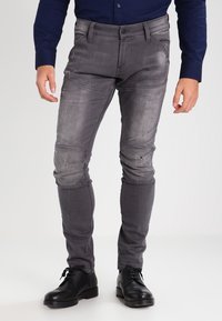Hombre vistiendo jeans ajustados grises desgastados, zapatos de cuero negro con cordones y una camisa de manga larga azul oscuro, de pie contra un fondo blanco.