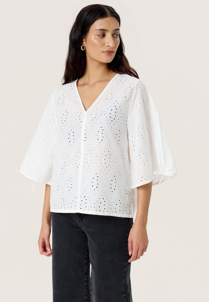 Blusa bianca con motivo ad occhiello, scollatura a V e maniche a sbuffo morbide, con dettagli a laccio ai polsini e una vestibilità rilassata.