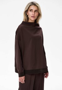 Vrouw met blond haar, die grote ringoorbellen draagt en een losse donkerbruine lange mouwtop en bijpassende broek aan heeft, kijkt opzij.