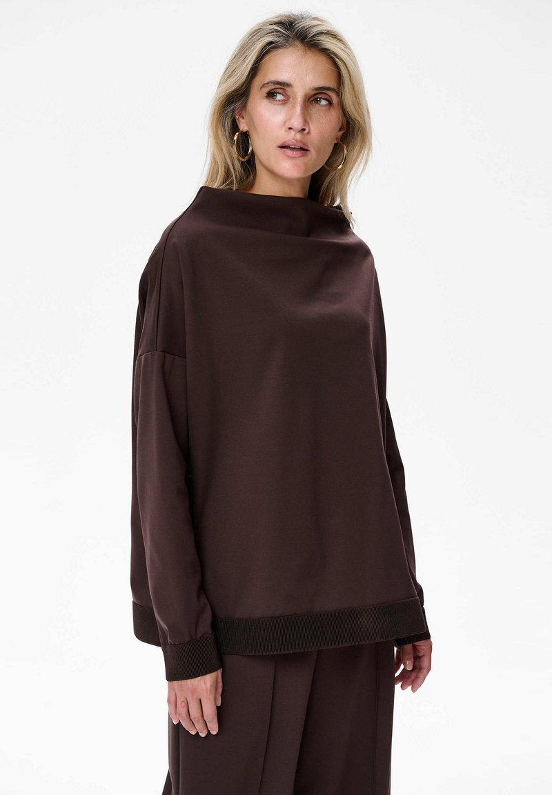 Vrouw met blond haar, die grote ringoorbellen draagt en een losse donkerbruine lange mouwtop en bijpassende broek aan heeft, kijkt opzij.