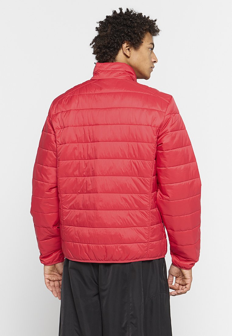 Chaqueta aislante roja con un diseño acolchado, cuello alto y mangas largas. Textura suave y material ligero. Pantalones negros visibles por debajo.