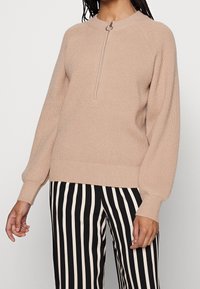 Beige knitted sweater with a half zip and ribbed texture, featuring raglan sleeves. Paired with black and white striped pants.