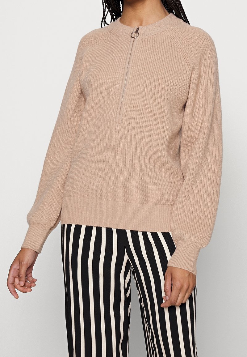 Beige knitted sweater with a half zip and ribbed texture, featuring raglan sleeves. Paired with black and white striped pants.
