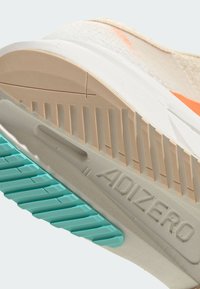 Ingrandimento di una suola di sneaker beige e arancione che mostra i battistrada color sabbia e teal e la scritta "ADIZERO" in rilievo sulla soletta bianca.