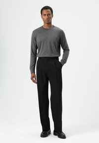Chemise grise à manches longues associée à un pantalon noir plissé. La tenue présente une coupe décontractée, une texture lisse et un design classique. Des chaussures noires complètent le look.
