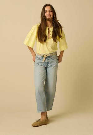 Jeune femme aux longs cheveux bruns portant un haut jaune, un jean bleu clair avec une ceinture beige et des chaussures marron, debout devant un fond beige.