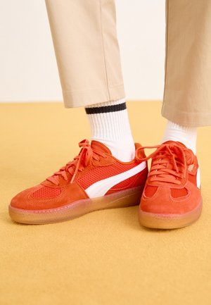 Osoba w beżowych spodniach, białych skarpetkach z czarnym paskiem oraz jaskrawo pomarańczowych trampkach z białymi paskami bocznymi stoi na żółtej podłodze.