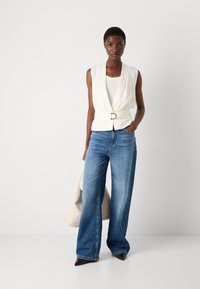 White sleeveless top with a draped front, belted at the waist; paired with wide-leg blue denim jeans and pointed black shoes.