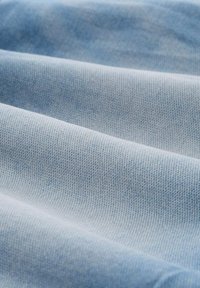 Hellblauer Denim-Stoff mit einer feinen, strukturierten Webart, die sanfte Falten und subtile Farbabweichungen auf der Oberfläche zeigt.